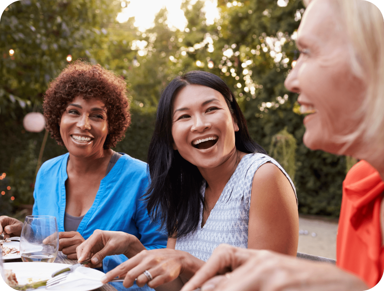 Three women smiling and laughing at an outdoor meal, showing off their natural looking dental implants.